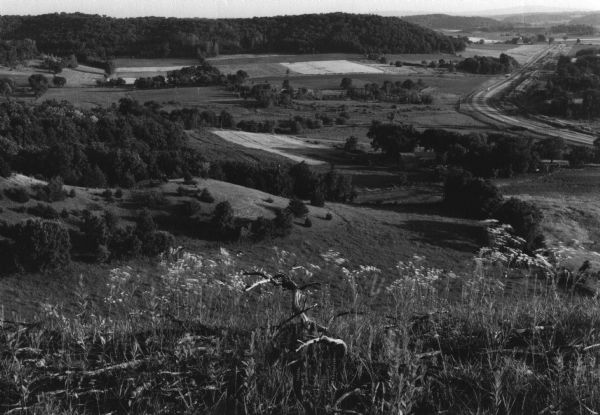 View from a bluff looking south. A highway is on the right; one farm building is among a stand of trees at the bottom of the hill.
