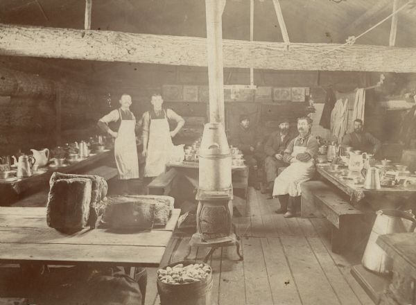 A group of six men in the dining hall of a lumber camp. The men are in a room with long tables filled with table settings.