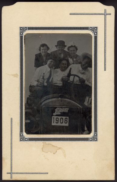 Tintype group portrait of three men and three women sitting in an open automobile. Two of the men are holding bottles in their hand. The sign on the front of the automobile reads: "Lambert," and a plate that says: "1906."