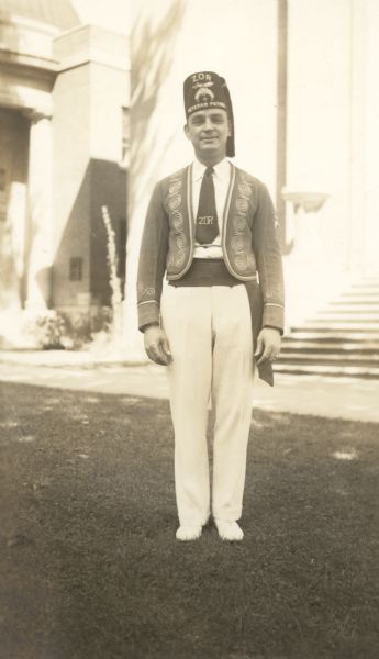 Outdoor portrait of an unidentified Shriner standing outdoors on a lawn. He is wearing a Zor Veteran Patrol fez, and a Zor necktie. The Zor Shrine is based in Madison and received its charter in 1933. There is a building in the background. 