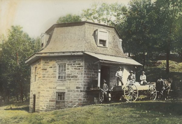 Group portrait of five men standing on and near a horse-drawn wagon in front of a grist mill, a stone building on the side of a hill. 