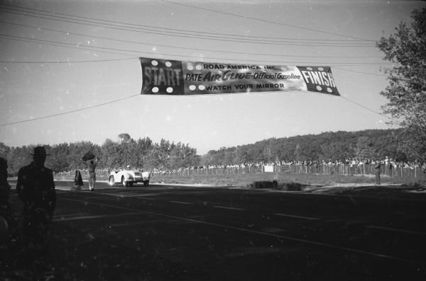 View towards a man driving an automobile towards a start and finish line. A banner over the course reads: "Start, Road America Inc., Pate Air Glide-Official Gasoline, Watch Your Mirror, Finish." Spectators are behind a fence on the other side of the course.