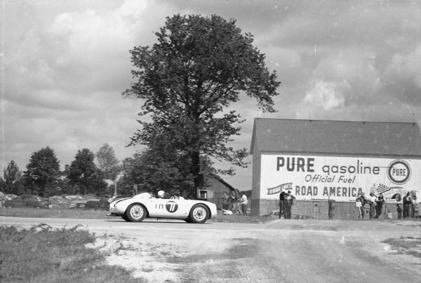 View from side of road towards the right side of a racecar being driven on a road. A barn on the other side of the road has a sign painted on it that reads: "Pure gasoline Office Fuel, Elkhart Lake's Road America." Spectators are watching from behind a fence.