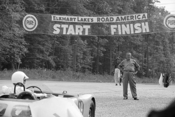 There is a man in a race car on the course in the foreground on the left. A man is standing on the road, under a banner that reads: "Pure, Elkhart Lake's Road America, Start, Finish."