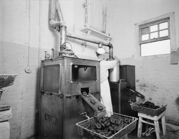 Gardner Baking Company making Purity Doughnuts. Doughnuts are coming down a chute from a machine, and a baker is standing and attending one of the machines.