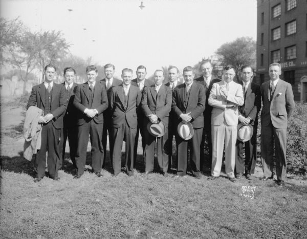 Group portrait of Gardner Baking Co. drivers/salesmen from out of town, taken outdoors near 849 E. Washington Avenue. Breese Stevens Field is in the background on the left, and on the right is the Simon Brothers Wholesale Grocery Warehouse, 901 E. Washington Avenue.