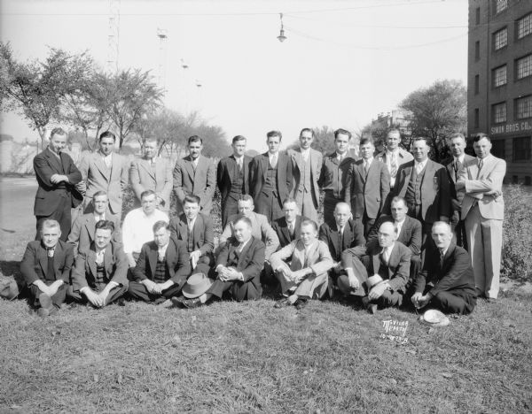 Group portrait of Gardner Baking Co. drivers/salesmen from Madison, taken outdoors near 849 E. Washington Avenue. Breese Stevens Field is in the background on the left, and on the right is the Simon Brothers Wholesale Grocery Warehouse, 901 E. Washington Avenue.