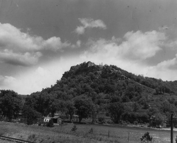 View of a wooded hill. Two small buildings, a fence, and railroad tracks are in the foreground. Caption reads: "Grandad Bluff, near La Crosse, Wis. This bluff towers over the city and has a lookout for tourists from which a great extent of the Mississippi River Valley can be seen."