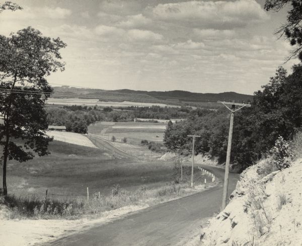 Slightly elevated view of a curving road, with telephone poles along it. The road leads down a hillside and is lined with fields and groves of trees. Caption reads: "Panorama Hy. 27 looking north between Northfield and Hixton, Jackson Co."