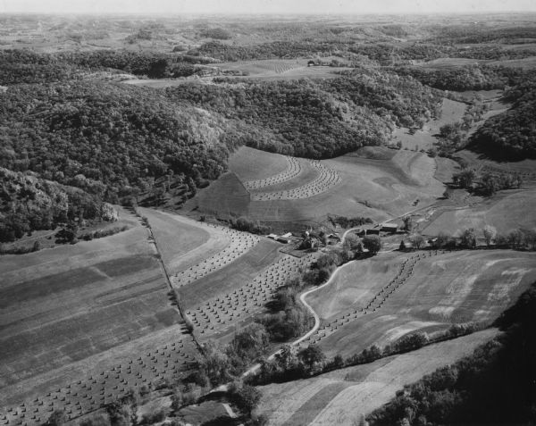 Aerial view of fields along hills, surrounded by forests. There are roads and buildings, and hay bales in the field.