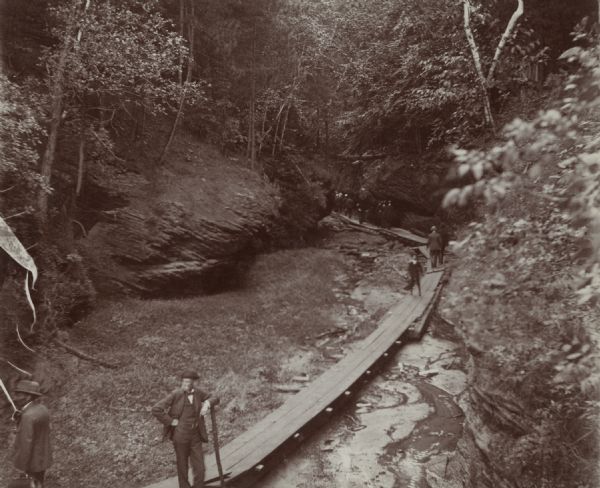 View of a man standing on a boardwalk in a ravine. Other people are walking along the boardwalk. A penciled note identifies the man as "RJ." Caption on rear of photograph reads: "Perhaps some day I will find a better picture of RJ which I will send you."