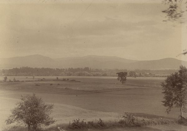 View across plains, with sparse trees. In the distance is a small body of water, with a forest and mountains in the distance.