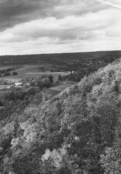 Elevated view along a forested hillside, overlooking farm fields, a barn, and a small town in the distance.