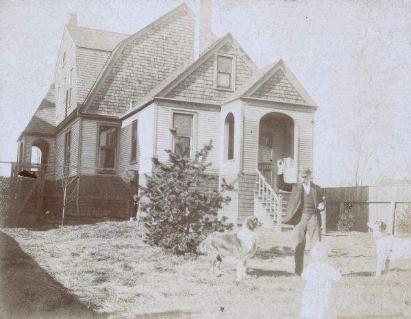 A man is standing in the yard of a house with two dogs and a young child. The man is holding up his hand, and the two dogs are looking up at something the man is holding. The child in the foreground has their back turned away from the camera.