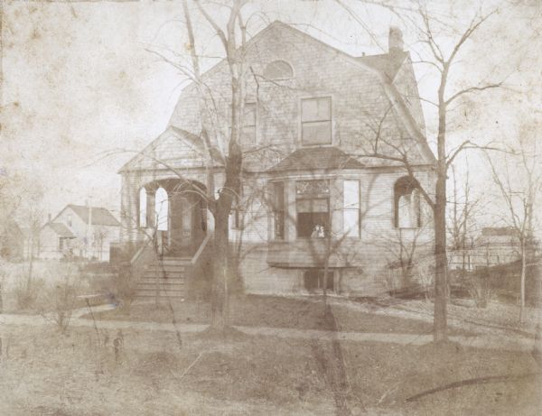 View towards a house, with a sidewalk in front, and a porch entrance on the left. A young child and a dog (probably Sigrid and Barry) are posing inside the front bay window.