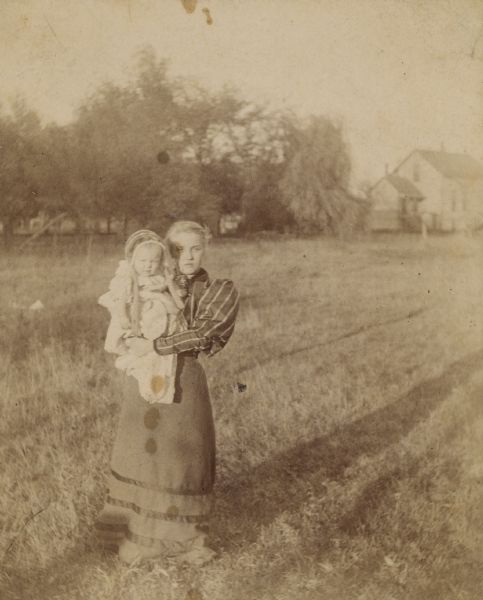 Outdoor portrait of a young woman holding an infant while standing in a field. There is a building near trees in the background.
