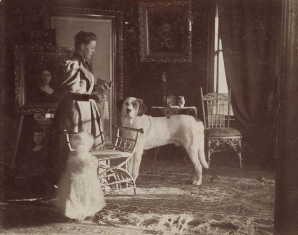 A large dog named Barry is standing in the center, and a woman, Sigrid's mother Hedwig, is standing behind a chair on the left. In front of the chair is a young child (Sigrid). Behind the group is a display of a number of framed portraits.