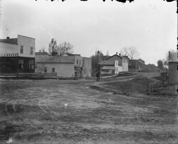 View of town, likely Iola, at an intersection. The street is unpaved. A man is riding a bicycle down the street heading towards another intersection in the background. A commercial building on the far left at the corner has a sign that reads: "[I]ola House." There are two more commercial buildings further down the street, and all have false fronts. Other buildings are in the background.