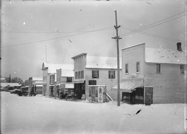 View down snowy street towards downtown Iola, including the Oddfellows Hall. Along the sidewalk is the Iola Hardware Store, and a Millinery Store. Advertising posters are posted on the sides of buildings, and on a large billboard along the sidewalk.