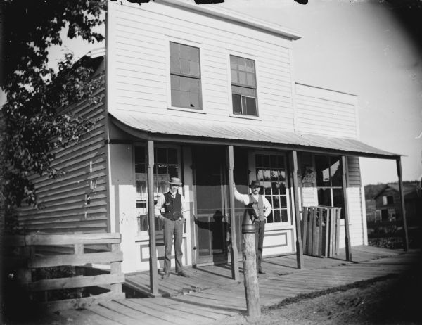 View from street towards two men, both wearing hats and vests, standing on the board sidewalk under the roofed entrance of a storefront. Advertisements for men's clothing is inside the window on the left. There is a horse-head hitching post along the unpaved street in the foreground.