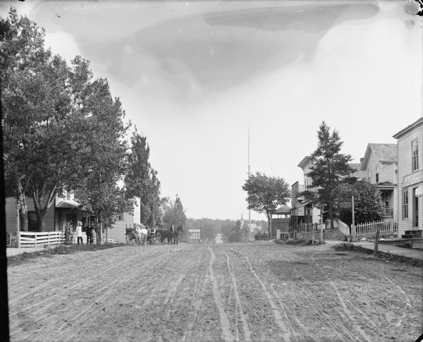 View down center of unpaved street. Commercial buildings are on both sides, and a group of men, one with a bicycle, are standing on the sidewalk on the left near a fence. In the street nearby, two men are sitting in a horse-drawn buggy, and a man behind them is sitting in a horse-drawn wagon. A dog is standing in the center of the street looking at the men. The street goes down a hill and continues towards trees in the distance, past a few houses and a church. In the foreground on the right is a building with a sign that reads, in part: "P.C. Gunderso_, Boot & Shoe Mar_." Further down the sidewalk is a sign on a post that reads: "D. Alfred Dale, Physician & Surgeon." Farther down the street is a scale with "Howe" painted on it. Behind the scale is a brick octagonal building with a roof, perhaps a pavilion, and near it is a tall pole, perhaps a flagpole. Also along the board sidewalks are hitching posts and lampposts.