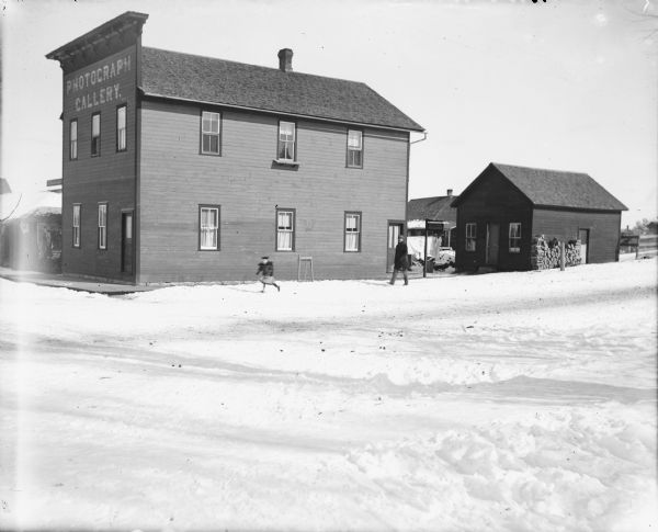 View across snowy street towards a two-story commercial building with a false front. The sign painted on the front reads "Photograph Gallery." A man and a young girl are walking in the snow along the side of the building. Another sign on a post behind the building reads: "W.R. Parks Photographer." There are other buildings behind the gallery, and one has a stack of fuel wood along the side. Laundry is handing to dry in the yard.