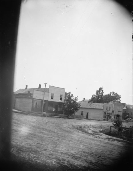 View towards intersection of unpaved street. People are sitting in front of the entrance of a commercial building at the corner on the left, and there is a sign (unreadable) hanging from a post at the curb. There are two more commercial buildings further down the street. The buildings all have false fronts.
