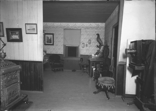 View inside what may be Parks's photography studio. There is a camera on the right, and a Sylvan Red Cross wood burning stove on the left. Through an open doorway is another room with a desk, chairs, and a taxidermy bird.