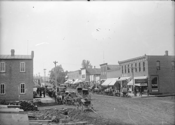 Slightly elevated view of downtown Iola. The curbs are lined with horse-drawn carriages and wagons, and people are gathered on the sidewalks in front of storefronts. In the foreground are stacks of lumber. On the street corner on the left is a brick buildings, and the sign on the awning reads: "_ardware." On the street corner on the right is the Thompson Hardware Co., next door is J. Ellandson, General Merchandise, and after that is the Bank of Iola. Many of the wagons are loaded up with milk cans.