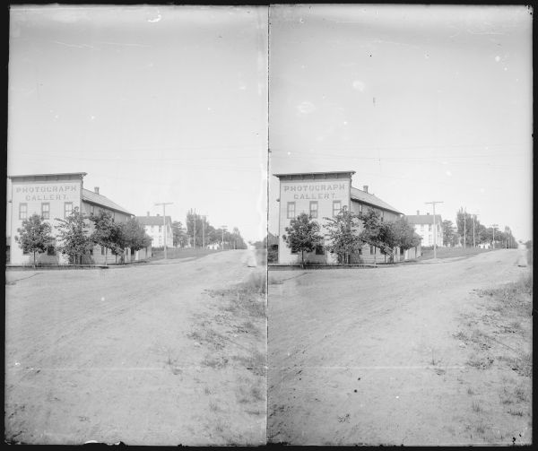 Exterior stereograph looking down street towards the Parks studio. The sign painted on the false front reads: "Photograph Gallery." A dog is standing near the side of the studio.