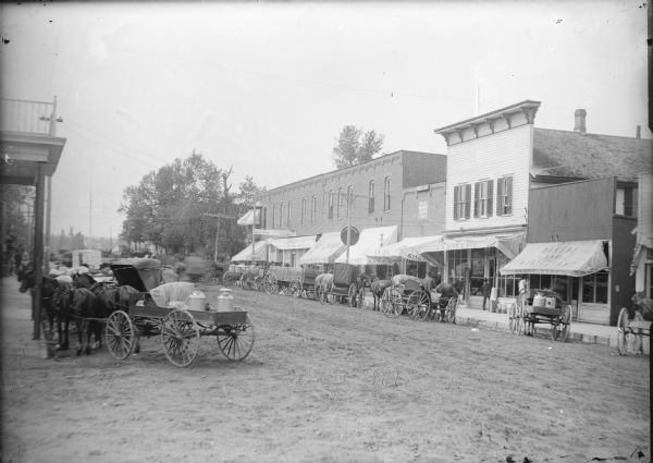 View looking down street towards storefronts. On the left side of the street, horse-drawn carriages and wagons are lined up at the curb, and more carriages and wagons are lined up across the street. Storefronts across the street include a Bakery and Restaurant; the H.M. Wipf & Bro. store; A. R Lea, Shoes; E.B. Benson Billiards & Pool; KROM Mercantile Company; and a bank. Written on negative at bottom center: "Main St., Iola, Wis."