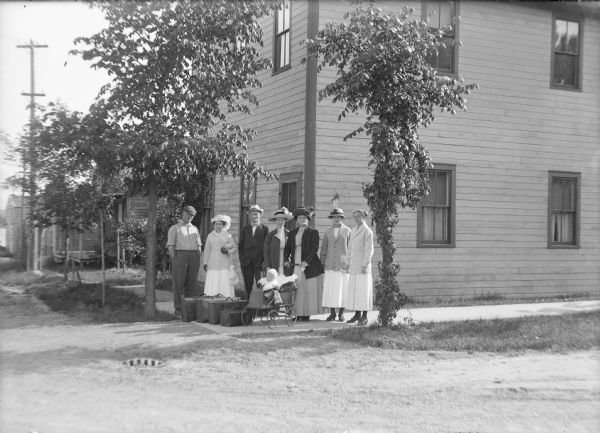 Group portrait of people, family and visitors, on the sidewalk in front of Parks House. Five women and two men are posing on the corner, and one woman is holding onto a baby carriage with an infant sitting inside. A row of luggage is on the sidewalk.