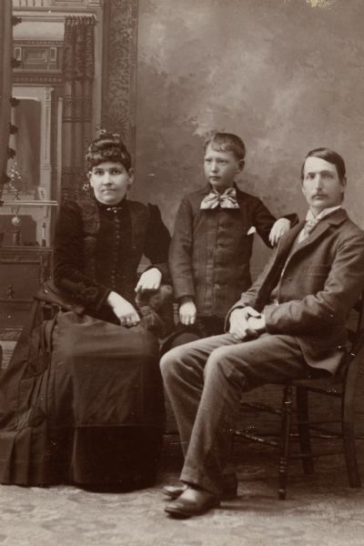 Studio portrait in front of a painted backdrop of a family, with a child standing between seated parents. These are Benjamin Woolsey Styles, his wife Elizabeth (David) Styles, and their youngest child, Edward.
