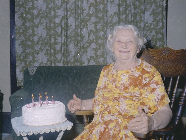 Emma Boyum, Sid's mother, is sitting in a rocking chair in a living room. A birthday cake with candles is on a small table in front of her.