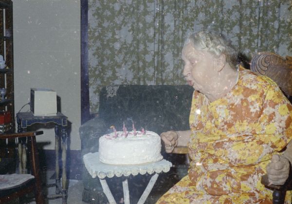 Emma Boyum, Sid's mother, is sitting in a rocking chair in a living room. She is blowing out the candles on a birthday cake which is on a small table in front of her.