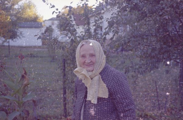 Waist-up view of Emma Boyum, Sid's mother, standing outdoors. She is wearing a scarf around her head. There are plants and trees behind her, and buildings are in the background.