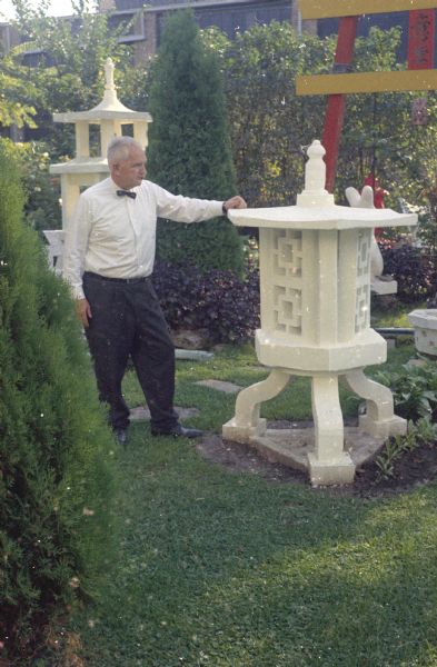 Sid is standing in his backyard posing with a sculpture. More sculptures and the torii gate are in the yard behind him among shrubs. The Madison-Kipp Corporation building is in the background.