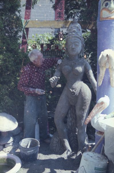 Sid is standing outdoors in his backyard working on a large sculpture  inspired by South Asian styles, which is shaded by trees and shrubs. There are other sculptures, including the torii gate, on the right and in the background. The Madison-Kipp Corporation building is in the far background.
