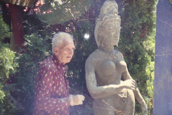 Waist-up view of Sid standing in his backyard next to a large sculpture inspired by South Asian styles. The yard is shaded by trees and shrubs. Part of another sculpture is in the background on the left.