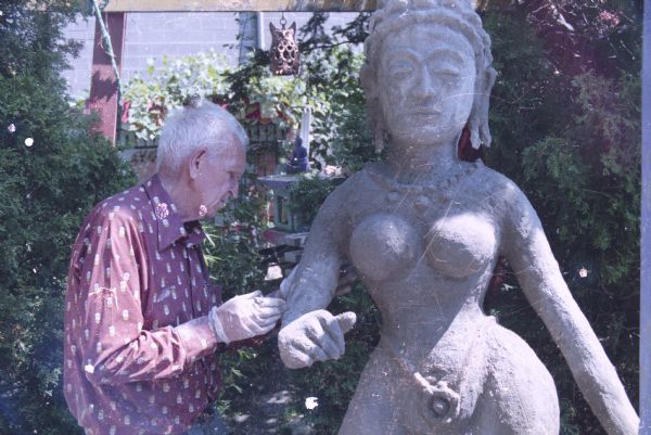 Waist-up view of Sid in his backyard working on a large sculpture  inspired by South Asian styles. There are shrubs and trees shading the yard. More sculptures are in the background, and the Madison-Kipp Corporation building is in the far background.