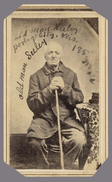 Carte-de-visite of a man wearing a long coat sitting in a chair next to a cloth covered table. He is holding a cane in front of him with both hands. Written on the print: "old man Seeley, Portage City, Wis. old man Seeley, 135 yrs. old."