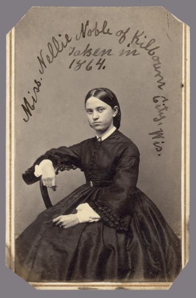 Carte-de-visite of Nellie Noble. She is sitting sideways in a chair. Written on print: "Miss Nellie Noble of Kilbourn City, Wis. Taken in 1864."