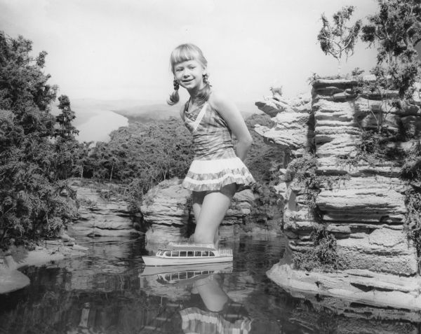 A young girl wearing a bathing suit is standing in almost knee-deep water in a Miniature model of the Wisconsin Dells. A small excursion boat is in the water in front of her. This completely landscaped exhibit was built to scale, 1/4 inch to a foot, faithfully depicting the Wisconsin Dells recreation area.