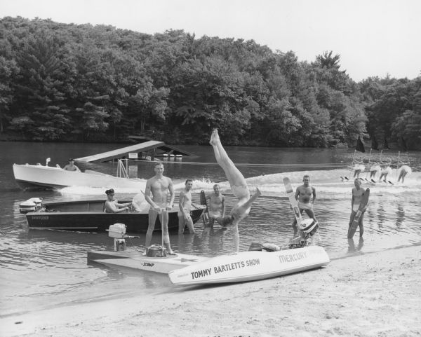 View towards group portrait of performers posing along the sandy shoreline. One man is doing a one-armed handstand on a motorboat in the foreground. Some of the men are holding water skis. In the background a group of four women are water skiing behind a boat, wearing costumes and holding flags. There is a ski jump further out in the lake, and a tree-lined shoreline is in the background.