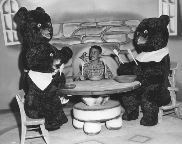 A young boy is sitting at a table with three stuffed bears, in a room decorated as if from the story of "Goldilocks and the Three Bears." The wall is painted with a mural that depicts a stone fireplace and two windows. 