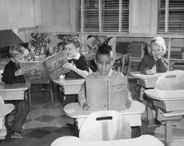 Four children are sitting at desks in a classroom. Two children are reading a book together on the left. A boy sitting in front is reading "Winnebago Boy," and a young girl is sitting and smiling at a desk on the right. The children are identified on the back of the print: Jacob Lone Tree, rear: John Schultz, Jackie Abrams, Debbie Reese.