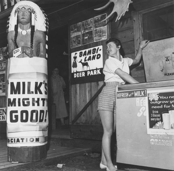 Patti Harrison is reaching into an open cooler in front of the Deer Park storefront. She is looking over her shoulder at a wooden American Indian advertising statue. There is a sign wrapped around the base of the carving that reads: "Milk's Mighty Good!" A sign on a door in the background reads: "Bambi-Land Deer Park."