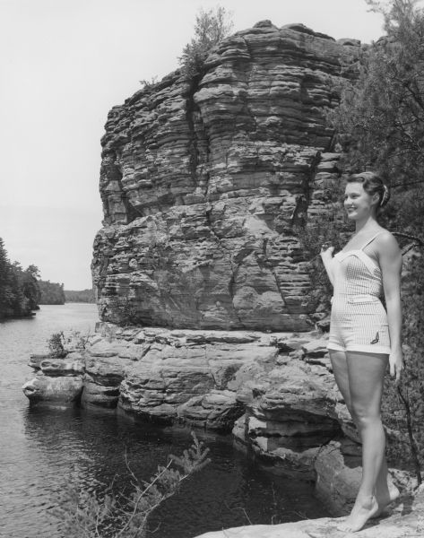 A young woman in a bathing suit is posing standing barefoot, and is holding onto the branch of a tree with her right hand. The Wisconsin River and a large rock formation are in the background.