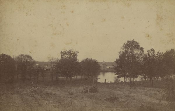 View across a field and Montello Lake. Buildings on the distant shore, seen through trees on the near shoreline, are the tower of the Montello House Hotel, and a smaller building on the right. Buffalo Lake is partially visible in the far background.