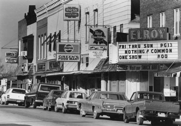 View along a commercial street in Elroy (now State Road 80). The Elroy Theater marquee is advertising screenings of the movie "Nothing in Common." Signs for commercial buildings include Ernie & Nita's Bar & Grill, Green's Food Mart, Shur Sav Food Stores, Fox Pharmacy, and Hansen's Clothing & Shoes. A Datsun truck is parked along the curb on the far right.